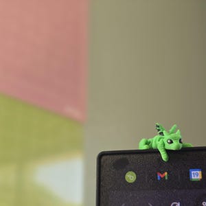 May include: A small, bright green dragon figurine with black-tipped wings and dark eyes, perched on the top edge of a black electronic screen. The dragon has a textured surface and is positioned to look as if it is resting on the frame of the device.