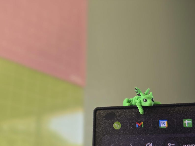 May include: A small, bright green dragon figurine with black-tipped wings and dark eyes, perched on the top edge of a black electronic screen. The dragon has a textured surface and is positioned to look as if it is resting on the frame of the device.