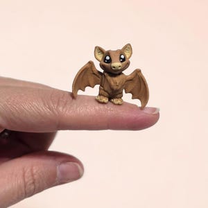 May include: A miniature brown bat figurine with large, expressive eyes and outstretched wings. The bat is perched on a fingertip, showcasing its small size and detailed craftsmanship. The figurine has a cute, cartoonish appearance.
