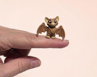 May include: A miniature brown bat figurine with large, expressive eyes and outstretched wings. The bat is perched on a fingertip, showcasing its small size and detailed craftsmanship. The figurine has a cute, cartoonish appearance.