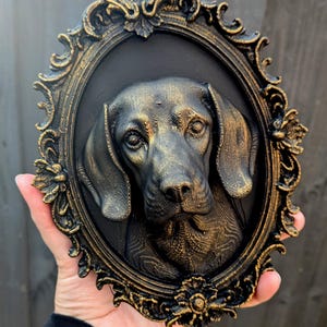 May include: A three-dimensional wall plaque featuring a sculpted dog head in a black and gold finish. The dog is centred within an ornate, oval-shaped frame that has intricate scrollwork details and a matching black and gold colour scheme.