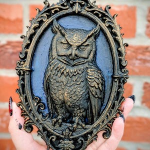 May include: An ornate, oval-shaped wall hanging featuring a detailed owl sculpture. The owl is rendered in shades of gold and brown against a blue background, framed by a black and gold filigree border. The piece is held against a brick wall.