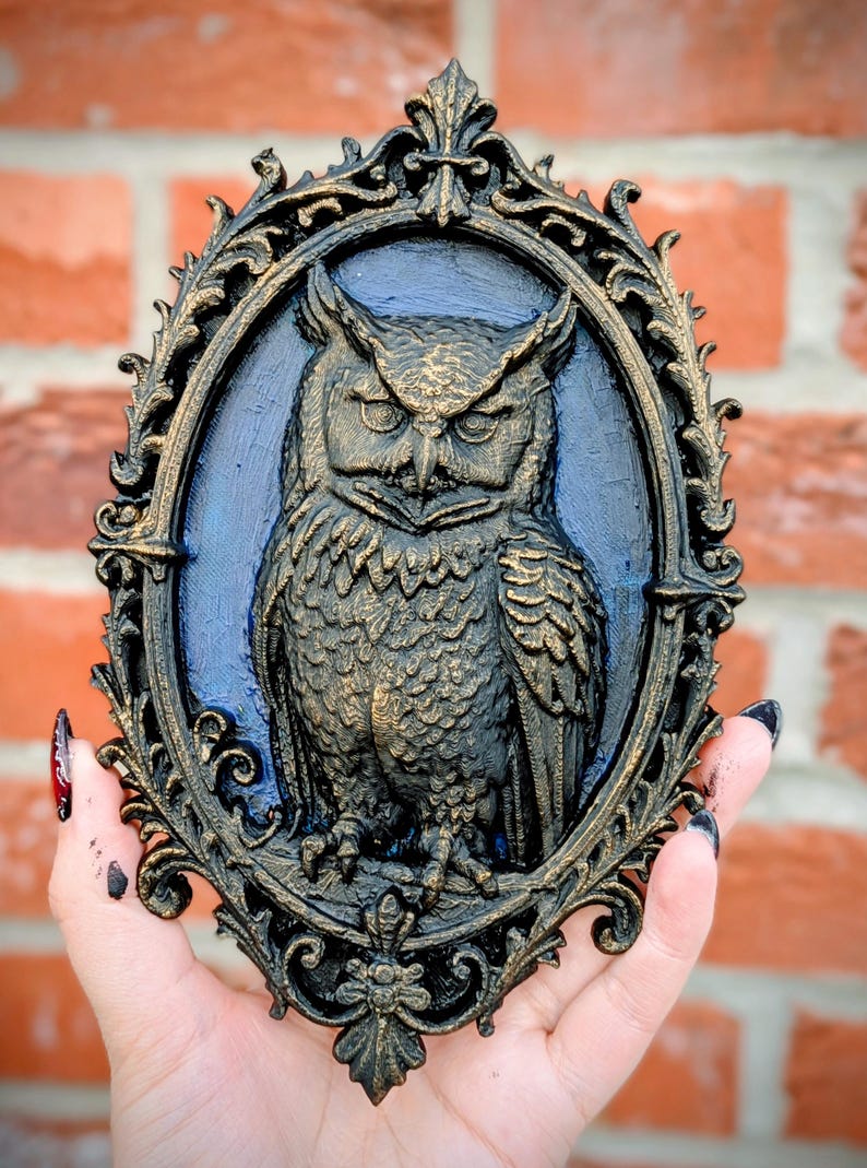 May include: An ornate, oval-shaped wall hanging featuring a detailed owl sculpture. The owl is rendered in shades of gold and brown against a blue background, framed by a black and gold filigree border. The piece is held against a brick wall.