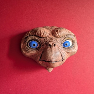 Könnte beinhalten: Detaillierte Skulptur von E.T.s Gesicht mit großen blauen Augen und strukturierter, hellbrauner Haut. Das Kunstwerk ist an einer roten Wand befestigt und erzeugt einen starken Farbkontrast.