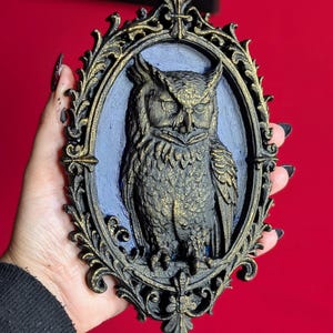 May include: An oval-shaped wall hanging featuring a detailed owl sculpture. The owl is dark with gold accents, set against a blue background within a decorative, ornate frame. The frame is black with gold highlights, creating a gothic aesthetic.
