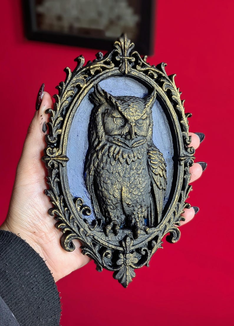 May include: An oval-shaped wall hanging featuring a detailed owl sculpture. The owl is dark with gold accents, set against a blue background within a decorative, ornate frame. The frame is black with gold highlights, creating a gothic aesthetic.