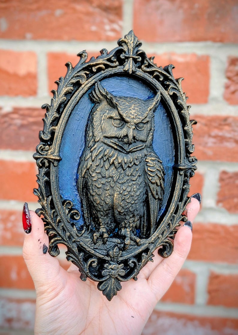 May include: Ornate, dark-colored wall decor featuring a detailed owl in an oval frame. The owl is depicted with intricate feather details and a stern expression, set against a blue background. The frame has a gold-colored trim.
