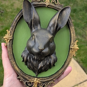 May include: A dark bronze rabbit head sculpture mounted on a green oval background, framed by an ornate gold-coloured border. The rabbit has long ears and detailed fur. The frame has decorative flourishes.