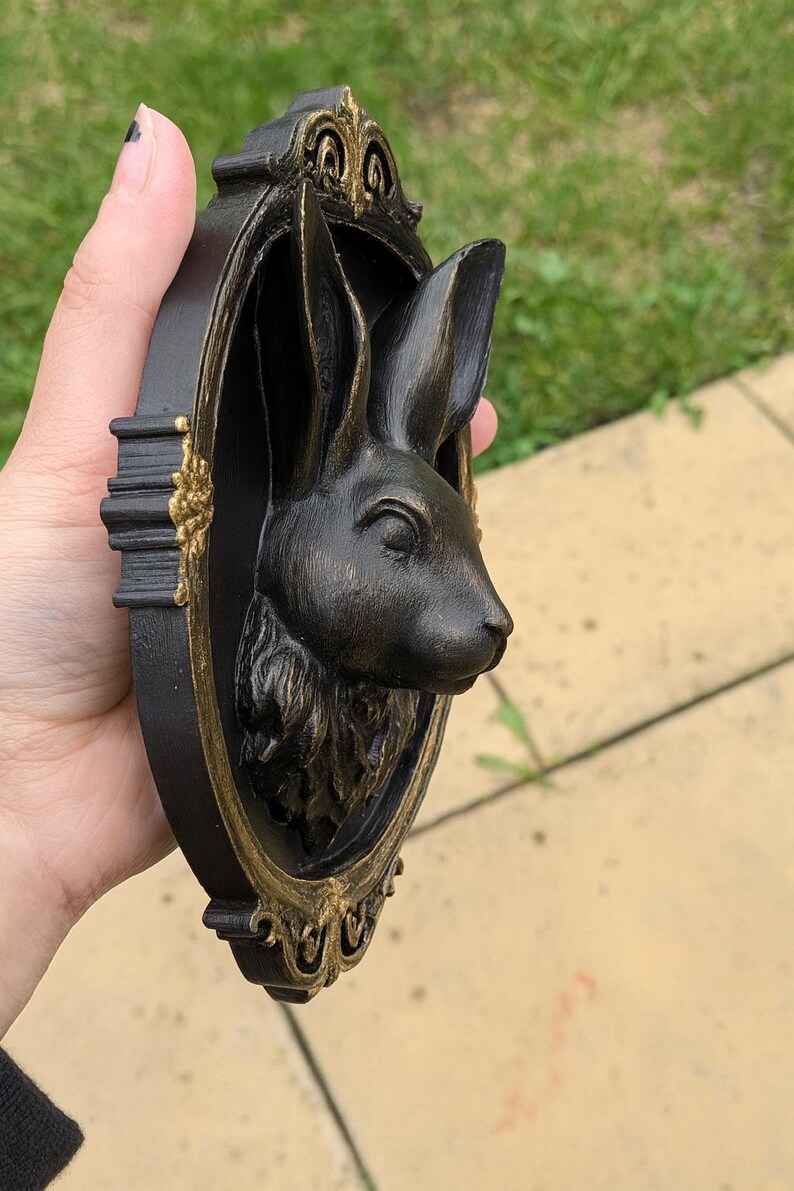Peut inclure: Sculpture murale t&ecirc;te de lapin noire et dor&eacute;e. La t&ecirc;te de lapin est dans un cadre ovale avec des d&eacute;tails dor&eacute;s. La sculpture est tenue dans une main sur un fond d'herbe verte et d'un patio en pierre.