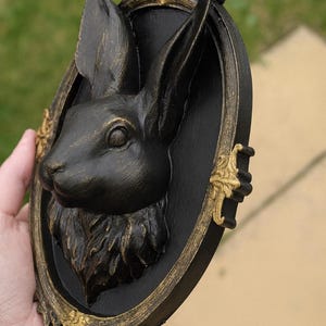 Peut inclure: D&eacute;coration murale t&ecirc;te de lapin noire dans un cadre ovale avec des accents dor&eacute;s. Le lapin est sculpt&eacute; avec des d&eacute;tails de fourrure et pr&eacute;sente une couronne royale. Le cadre a des d&eacute;tails dor&eacute;s orn&eacute;s, cr&eacute;ant une esth&eacute;tique vintage.