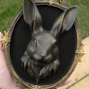 Peut inclure: Sculpture de t&ecirc;te de lapin couleur bronze avec de longues oreilles, sur fond ovale noir. La sculpture est encadr&eacute;e d'une bordure dor&eacute;e orn&eacute;e. L'&oelig;uvre est une d&eacute;coration murale.