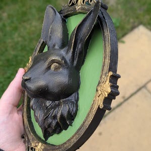 May include: A black rabbit head sculpture mounted on a green oval background, framed in a black and gold ornate border. The rabbit has detailed fur and is set against a green backdrop, creating a striking contrast. The frame has decorative gold accents.