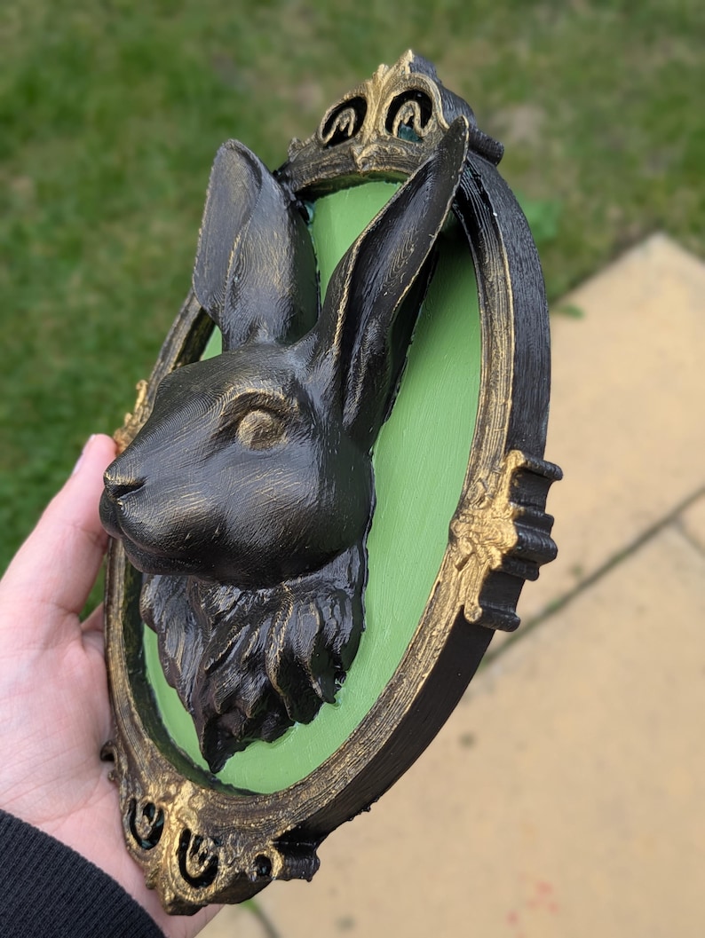 May include: A black rabbit head sculpture mounted on a green oval background, framed in a black and gold ornate border. The rabbit has detailed fur and is set against a green backdrop, creating a striking contrast. The frame has decorative gold accents.