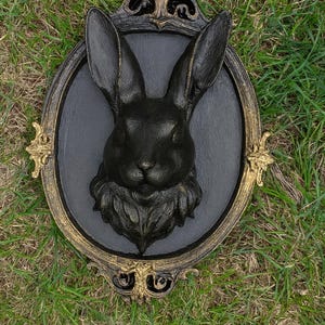 Peut inclure: D&eacute;coration murale t&ecirc;te de lapin noire avec un cadre dor&eacute; et noir orn&eacute;. Le lapin est centr&eacute; sur un ovale gris fonc&eacute;, avec des d&eacute;tails de fourrure et des traits du visage. Le cadre a des ornements d&eacute;coratifs.