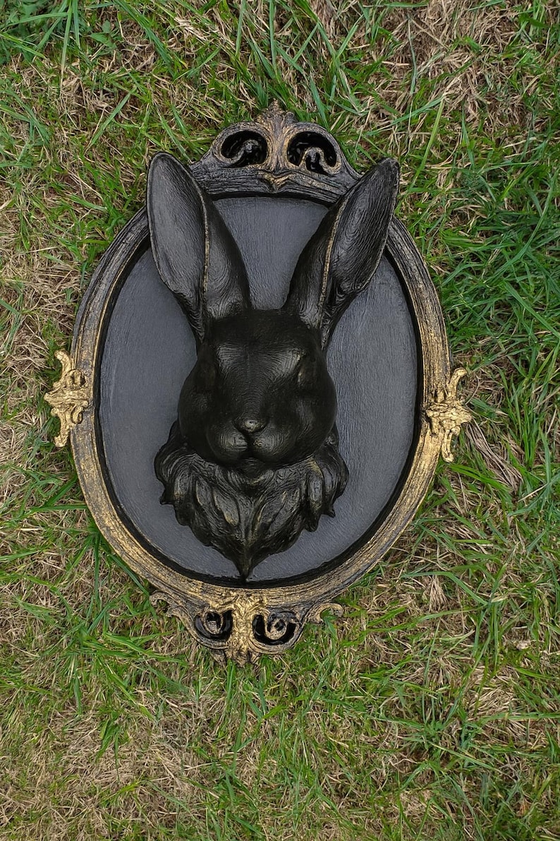 Peut inclure: D&eacute;coration murale t&ecirc;te de lapin noire avec un cadre dor&eacute; et noir orn&eacute;. Le lapin est centr&eacute; sur un ovale gris fonc&eacute;, avec des d&eacute;tails de fourrure et des traits du visage. Le cadre a des ornements d&eacute;coratifs.