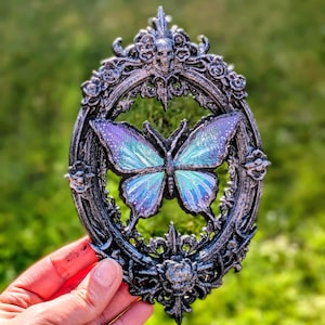 May include: A decorative, oval-shaped wall hanging featuring a butterfly with iridescent blue and purple wings. The butterfly is set within a detailed, ornate frame with a skull at the top and floral accents. The frame has a dark, metallic finish.