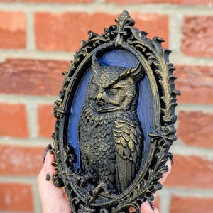May include: A black and gold owl relief sculpture in an ornate oval frame. The owl is detailed with textured feathers and sits against a blue background. The frame has a decorative, scrollwork design.