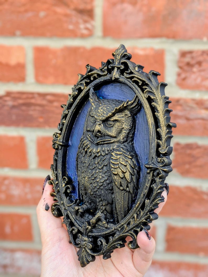 May include: A black and gold owl relief sculpture in an ornate oval frame. The owl is detailed with textured feathers and sits against a blue background. The frame has a decorative, scrollwork design.