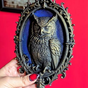 May include: An ornate, oval-shaped wall hanging featuring a detailed relief of an owl. The owl is depicted in shades of gold and black against a blue background, framed by a decorative, dark gold border. The piece has a vintage, gothic aesthetic.