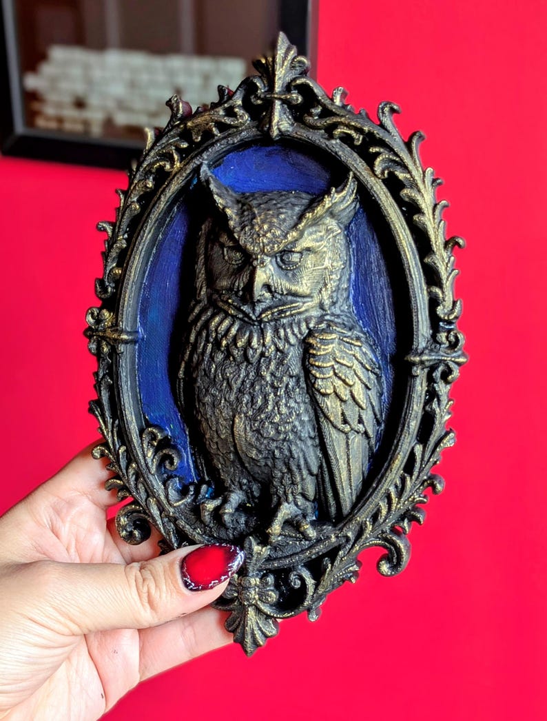 May include: An ornate, oval-shaped wall hanging featuring a detailed relief of an owl. The owl is depicted in shades of gold and black against a blue background, framed by a decorative, dark gold border. The piece has a vintage, gothic aesthetic.