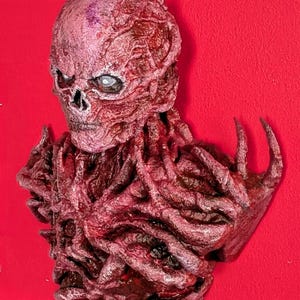 May include: A detailed, reddish-brown bust of a creature with a skull-like face and textured, organic features. The sculpture has a menacing appearance with visible veins and a rough, weathered texture. The background is a solid, vibrant red.