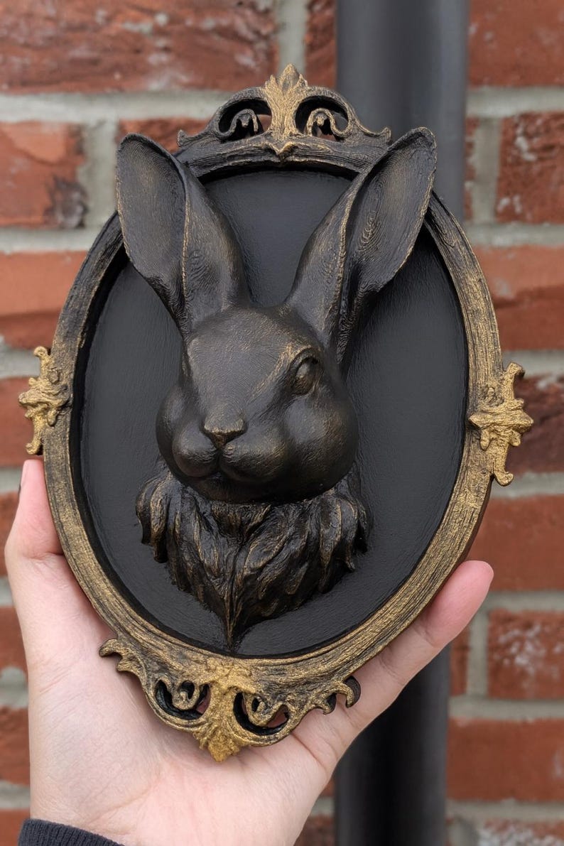 Peut inclure: Sculpture de t&ecirc;te de lapin couleur bronze, mont&eacute;e sur une plaque ovale noire avec un cadre dor&eacute; orn&eacute;. Le lapin a de longues oreilles et une fourrure d&eacute;taill&eacute;e. Le cadre pr&eacute;sente des ornements d&eacute;coratifs.