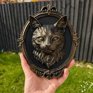 May include: A three-dimensional wall plaque featuring a sculpted cat head in a metallic gold finish, set against a black oval background. The cat has detailed fur textures and pointed ears. The piece is encased in an ornate, decorative gold frame.