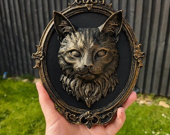 May include: A three-dimensional wall plaque featuring a sculpted cat head in a metallic gold finish, set against a black oval background. The cat has detailed fur textures and pointed ears. The piece is encased in an ornate, decorative gold frame.