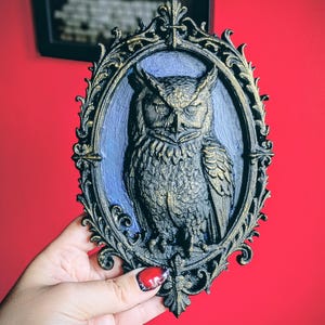 May include: An ornate, oval-shaped wall plaque featuring a detailed relief of an owl. The owl is depicted in shades of gold and black, set against a blue background within a decorative frame. The frame has a black base with gold accents.