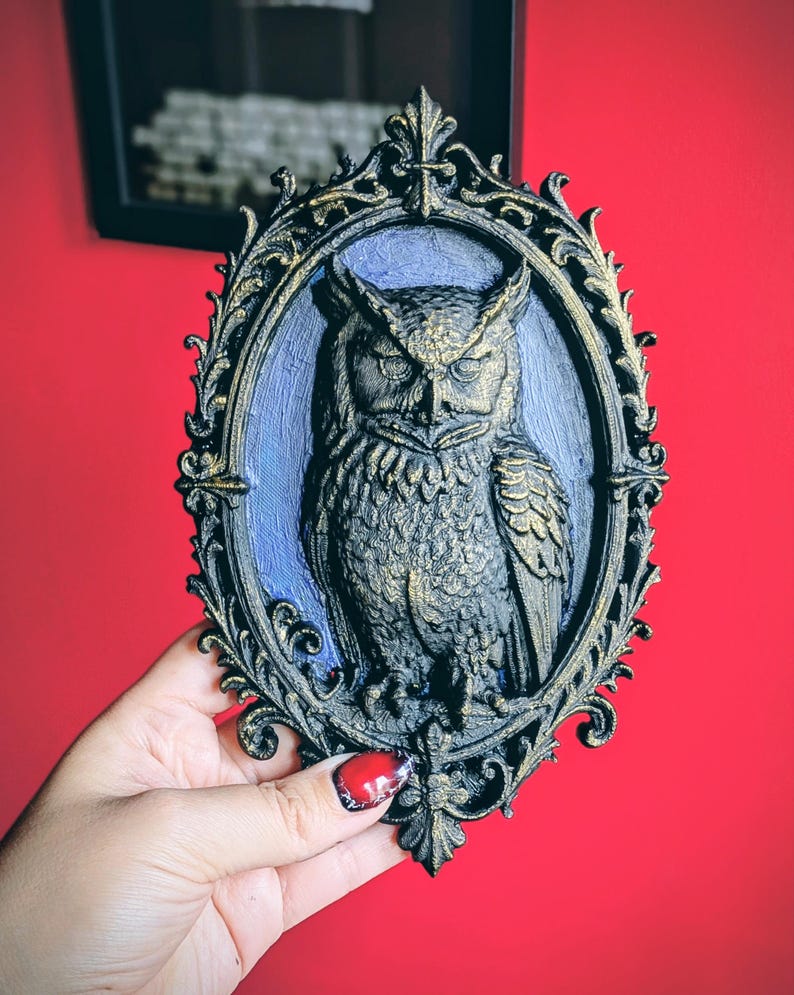 May include: An ornate, oval-shaped wall plaque featuring a detailed relief of an owl. The owl is depicted in shades of gold and black, set against a blue background within a decorative frame. The frame has a black base with gold accents.