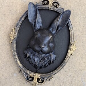 Peut inclure: D&eacute;coration murale t&ecirc;te de lapin noire dans un cadre ovale avec des accents dor&eacute;s. Le lapin est centr&eacute; sur un fond noir, avec de longues oreilles et une texture de fourrure d&eacute;taill&eacute;e. Le cadre a des d&eacute;tails orn&eacute;s.