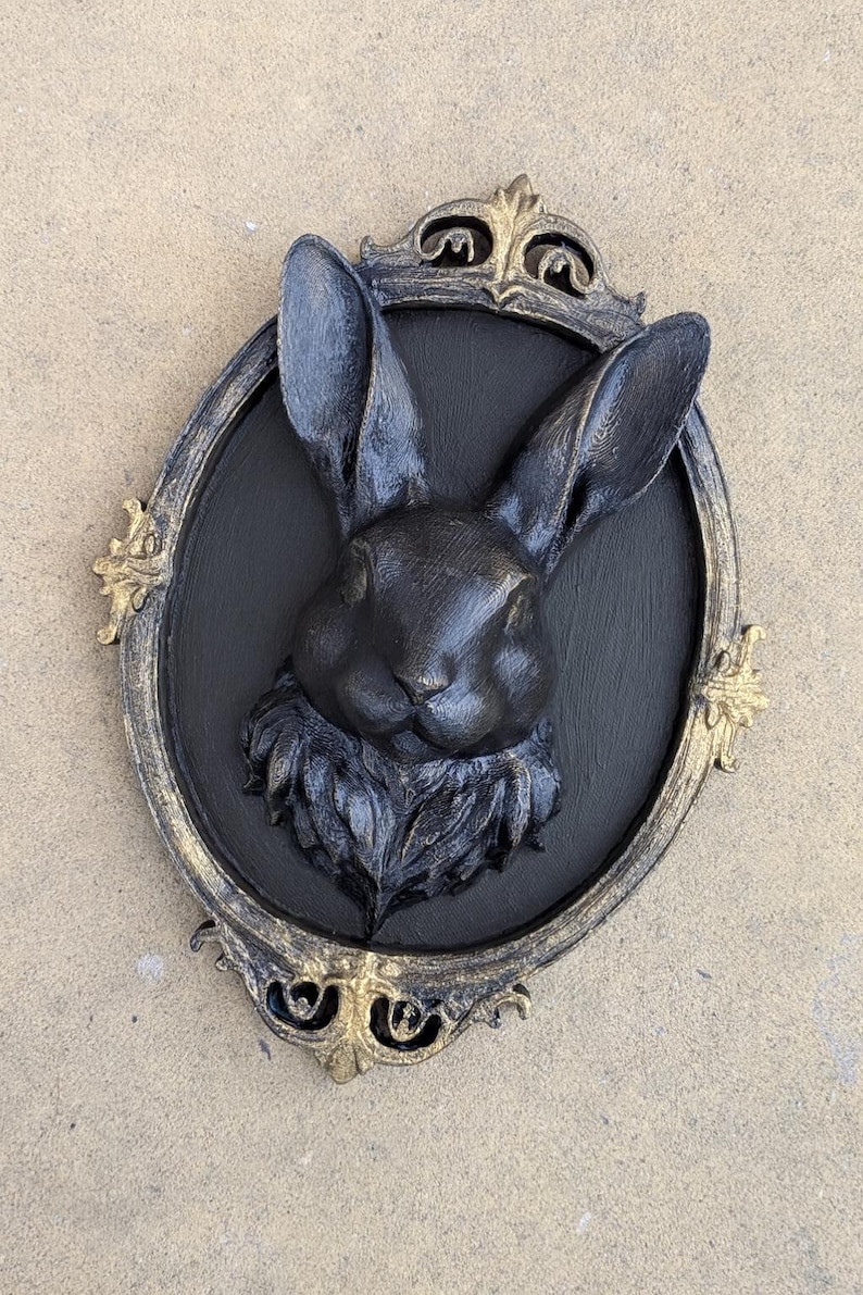 Peut inclure: D&eacute;coration murale t&ecirc;te de lapin noire dans un cadre ovale avec des accents dor&eacute;s. Le lapin est centr&eacute; sur un fond noir, avec de longues oreilles et une texture de fourrure d&eacute;taill&eacute;e. Le cadre a des d&eacute;tails orn&eacute;s.