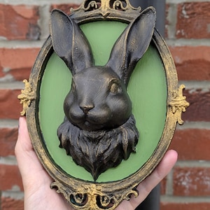May include: A bronze-colored rabbit head sculpture mounted on a green oval background, framed with ornate gold detailing. The sculpture is held in a hand, showcasing the detailed texture of the rabbit's fur and the decorative frame.