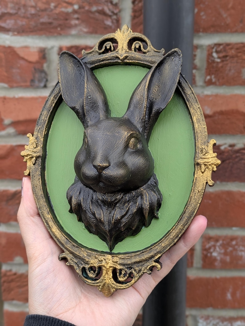 May include: A bronze-colored rabbit head sculpture mounted on a green oval background, framed with ornate gold detailing. The sculpture is held in a hand, showcasing the detailed texture of the rabbit's fur and the decorative frame.