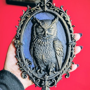 May include: A detailed, oval-shaped wall hanging featuring a sculpted owl in a decorative frame. The owl is dark gray with gold accents, set against a blue background. The frame is black with gold trim. The overall design has a gothic or vintage aesthetic.