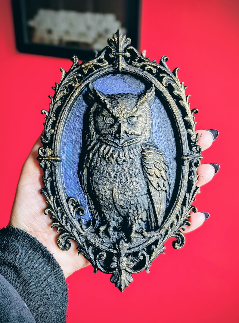May include: A detailed, oval-shaped wall hanging featuring a sculpted owl in a decorative frame. The owl is dark gray with gold accents, set against a blue background. The frame is black with gold trim. The overall design has a gothic or vintage aesthetic.