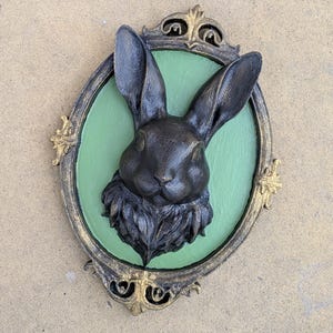 May include: A dark bronze-colored rabbit head sculpture mounted on a green oval background, framed by an ornate gold and black border. The rabbit has detailed fur and long ears, creating a vintage aesthetic.