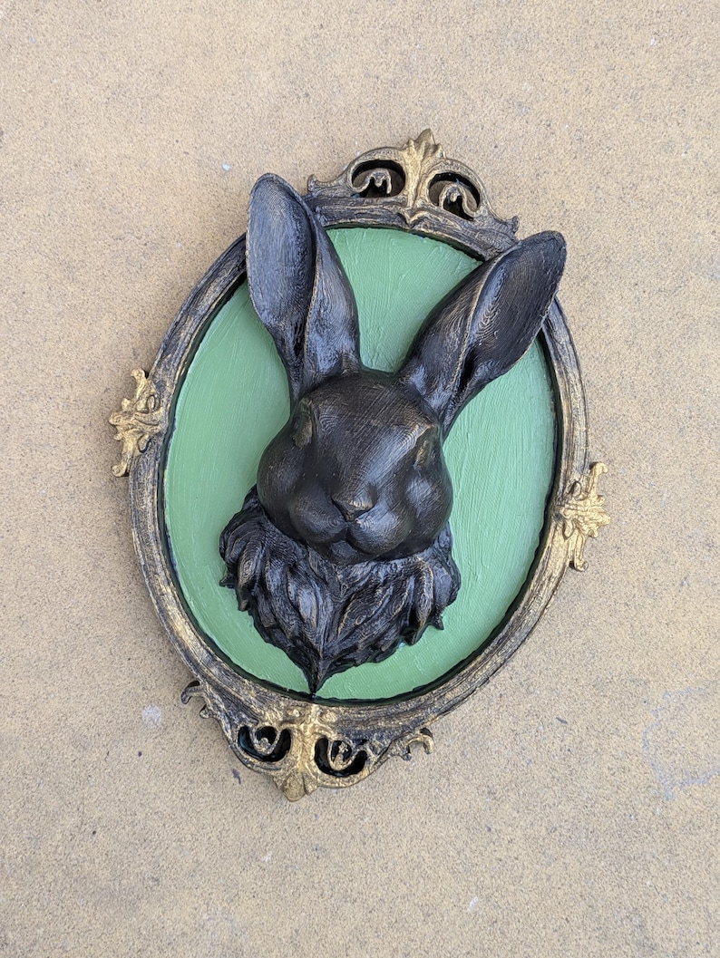 May include: A dark bronze-colored rabbit head sculpture mounted on a green oval background, framed by an ornate gold and black border. The rabbit has detailed fur and long ears, creating a vintage aesthetic.