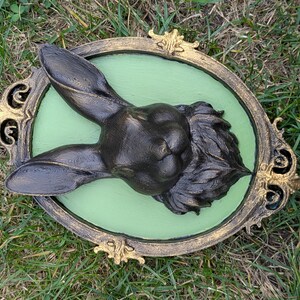 May include: A dark bronze rabbit head sculpture mounted on a light green oval background, framed by a decorative gold and black ornate border. The rabbit's ears are upright, and the fur is detailed.