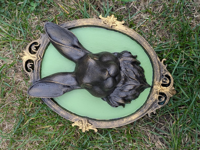 May include: A dark bronze rabbit head sculpture mounted on a light green oval background, framed by a decorative gold and black ornate border. The rabbit's ears are upright, and the fur is detailed.