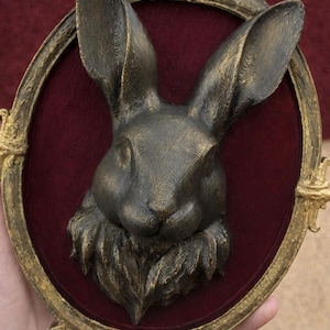 May include: A bronze-colored rabbit bust mounted on a burgundy oval background, framed by an ornate gold-colored border. The rabbit has detailed fur texture and long ears, creating a vintage aesthetic.