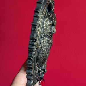 May include: A black and gold owl-shaped wall hanging. The owl is intricately detailed with a textured surface and a decorative frame. The artwork is set against a red background, highlighting the sculpture's dark colors and ornate design.