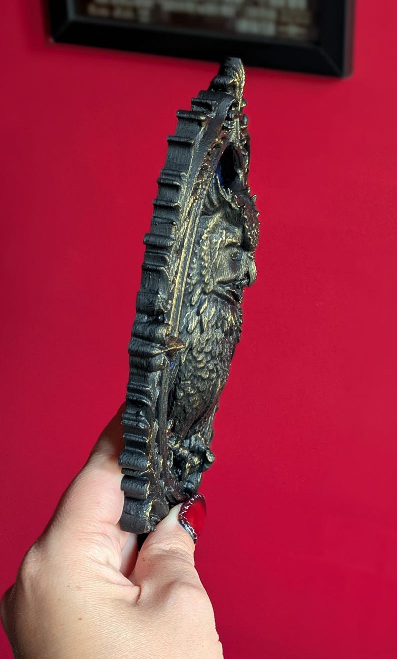 May include: A black and gold owl-shaped wall hanging. The owl is intricately detailed with a textured surface and a decorative frame. The artwork is set against a red background, highlighting the sculpture's dark colors and ornate design.
