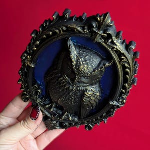 May include: A black and gold owl relief sculpture in an ornate frame. The owl is centered on a blue background. The frame has a detailed, vintage design. The sculpture is held against a red background.