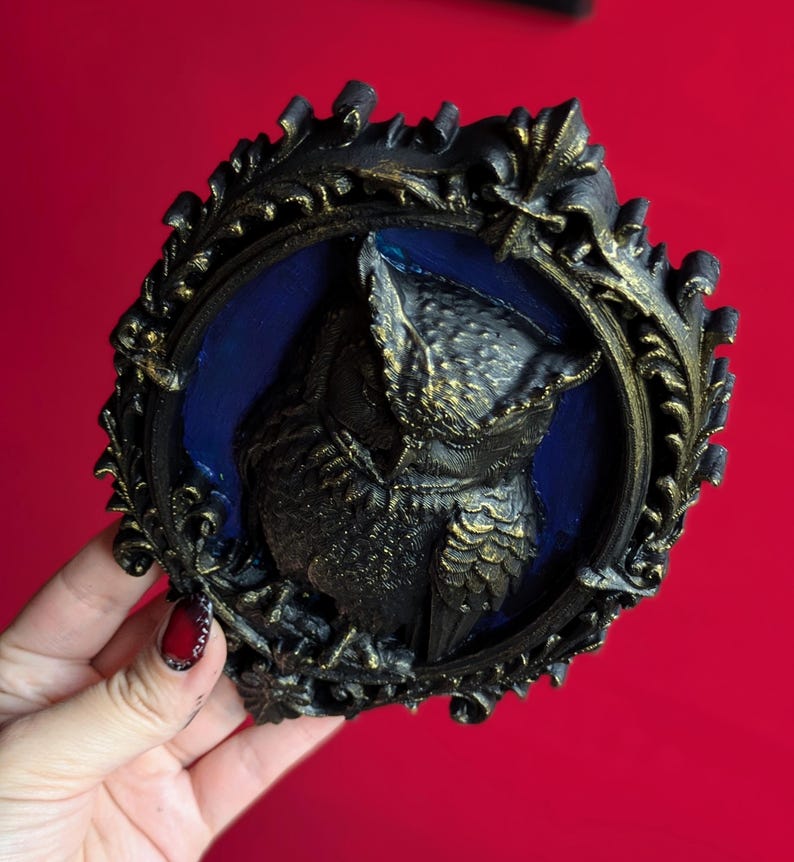 May include: A black and gold owl relief sculpture in an ornate frame. The owl is centered on a blue background. The frame has a detailed, vintage design. The sculpture is held against a red background.