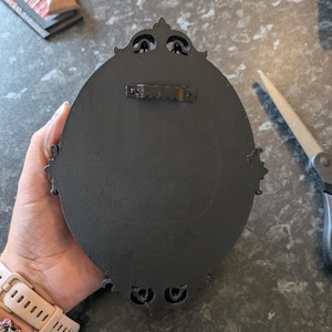 Peut inclure: D&eacute;coration murale ovale noire avec des d&eacute;tails orn&eacute;s. Le dos comporte un crochet en dents de scie pour le montage. Le cadre pr&eacute;sente des &eacute;l&eacute;ments d&eacute;coratifs en haut et en bas. La d&eacute;coration est tenue par une main.