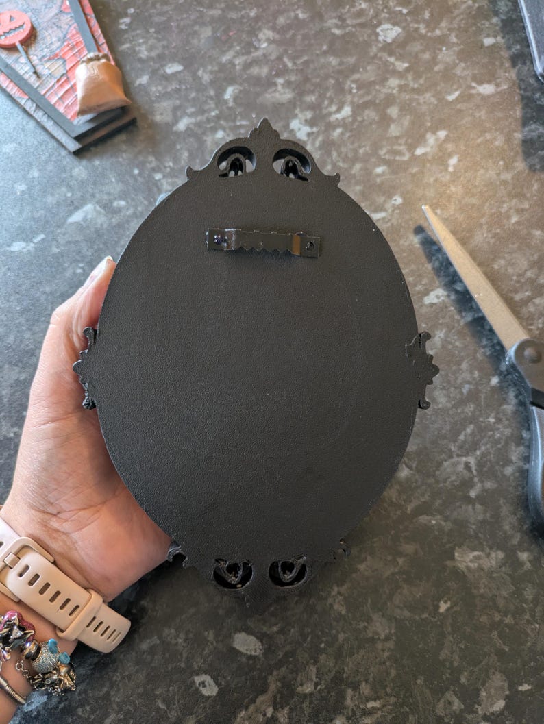 Peut inclure: D&eacute;coration murale ovale noire avec des d&eacute;tails orn&eacute;s. Le dos comporte un crochet en dents de scie pour le montage. Le cadre pr&eacute;sente des &eacute;l&eacute;ments d&eacute;coratifs en haut et en bas. La d&eacute;coration est tenue par une main.
