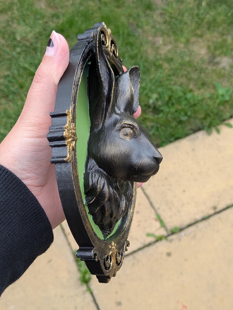 May include: Black rabbit head wall decor with a gold and black frame. The rabbit head is in profile, set against a green background. The frame has ornate gold detailing.