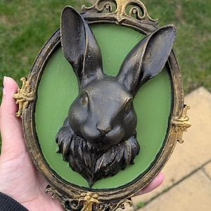 May include: A dark gray rabbit head sculpture with long ears, set against a green background within an ornate, oval, gold-trimmed frame. The frame has decorative flourishes. The sculpture is held in a hand.