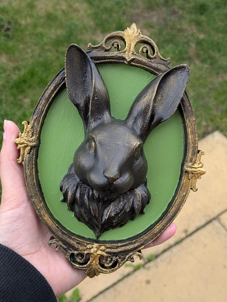 May include: A dark gray rabbit head sculpture with long ears, set against a green background within an ornate, oval, gold-trimmed frame. The frame has decorative flourishes. The sculpture is held in a hand.